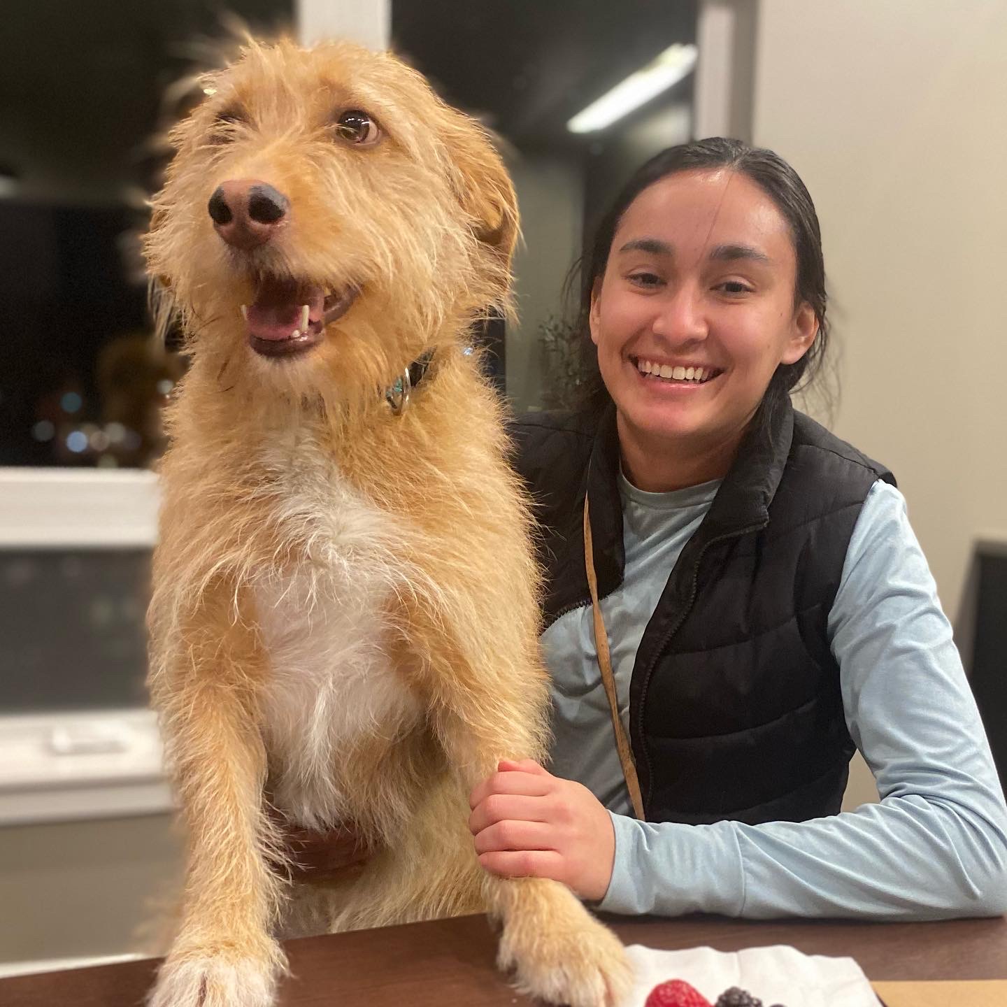 Image of Sami sitting at a table with her fluffy village dog Rafa in her lap with front legs on the table smiling
